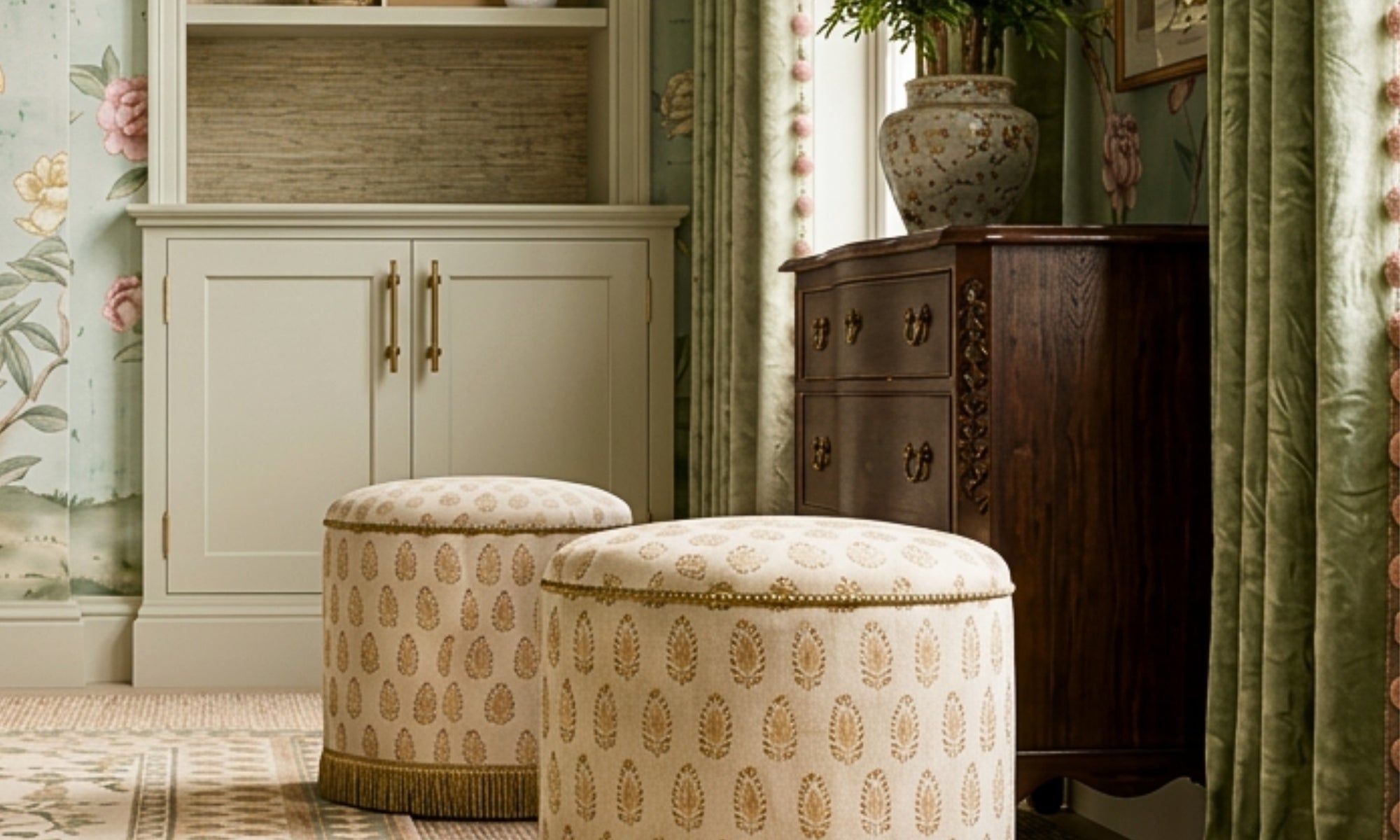 Two round upholstered ottomans with a beige and gold botanical pattern, styled in a classic room setting next to a dark wood cabinet and green curtains.