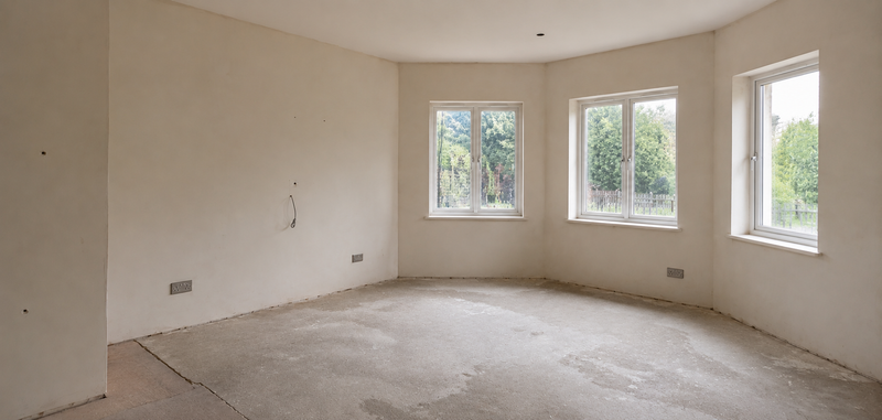 An empty room with unfinished concrete floors and off-white walls, featuring three double-paned windows looking out onto green trees.