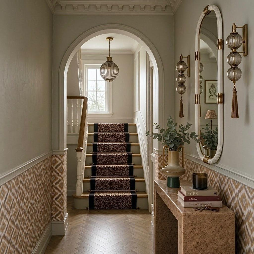 An elegant interior hallway of a Victorian renovation with a staircase covered in a dark patterned runner rug, flanked by a large oval wall mirror and decorative wall sconces. The space features warm beige walls, herringbone wood flooring, and a marble console table with books and a vase.