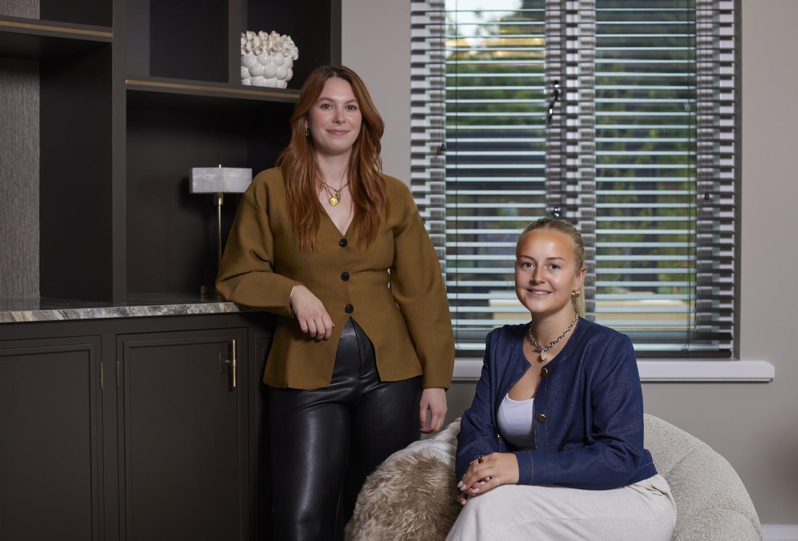 Two women sitting in a room with a window and shelves in the background
