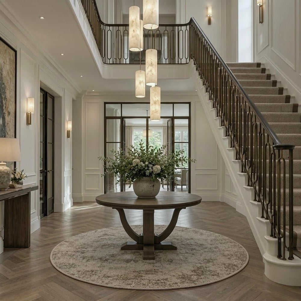 A grand two-story entryway with a round wooden entry table on a distressed beige rug, a large floral arrangement, a sweeping staircase with dark wood railings, and elegant cylindrical pendant lights hanging from the ceiling.